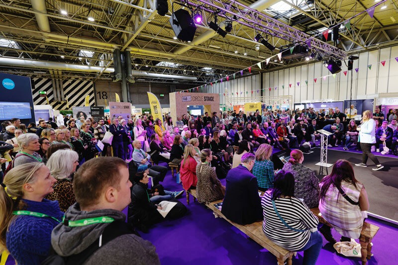 A large crowd gathers in a purple and white room at the FAB UK show, showcasing a vibrant atmosphere and engagement.