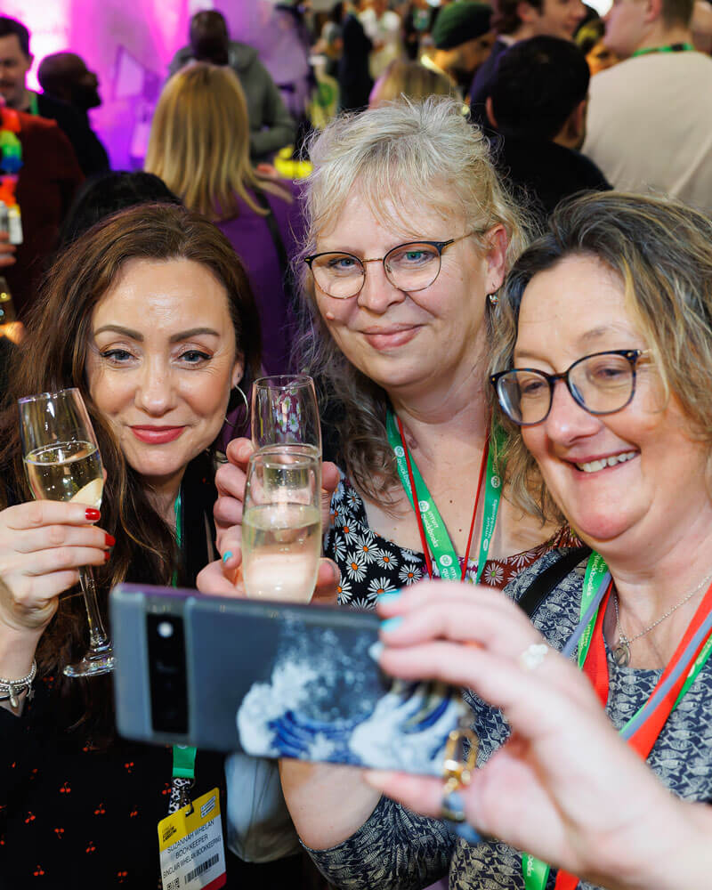 A woman holding a cell phone, taking a selfie with friends at the FAB UK show.